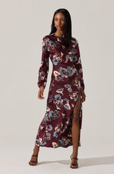 A woman is wearing a long, floral dress with a high slit. The dress has a maroon background with large, abstract floral patterns in shades of blue, beige, and tan. It features long sleeves with gathered cuffs and a round neckline.